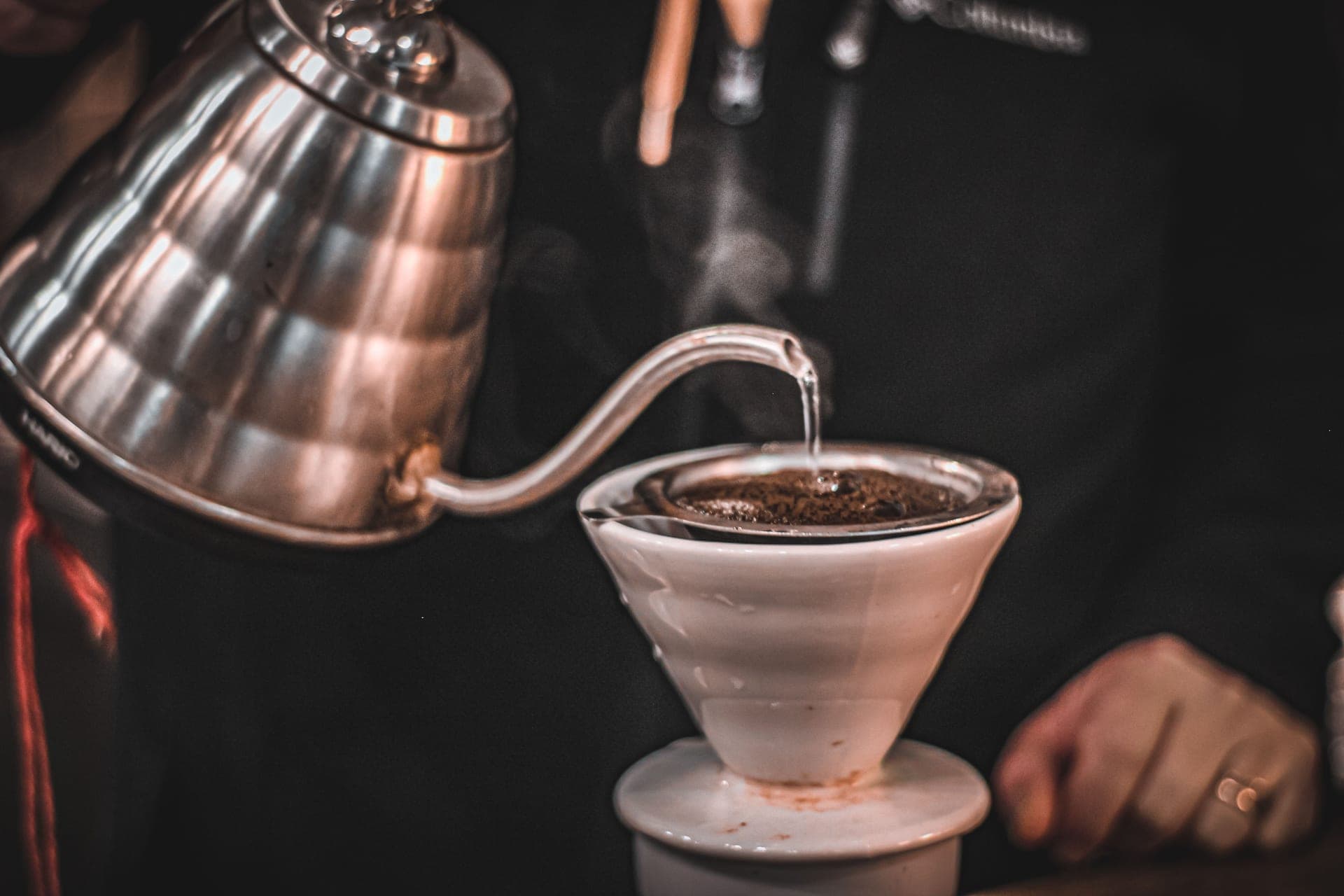 A gooseneck kettle pouring water over a coffee dripper on a dark background